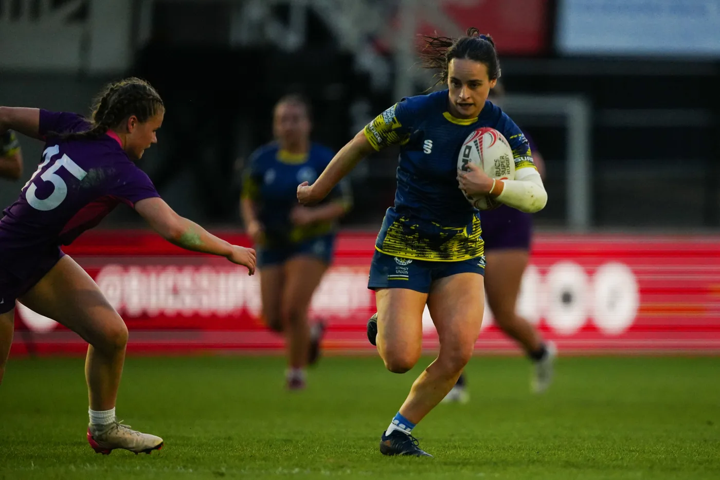 A player in navy and yellow runs with the ball as an opponent in purple dives to tackle