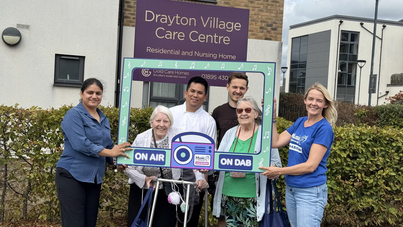 Residents and staff outside the Drayton Village Care Centre sign with the Music for Dementia team, holding an "On air, on DAB" photo frame