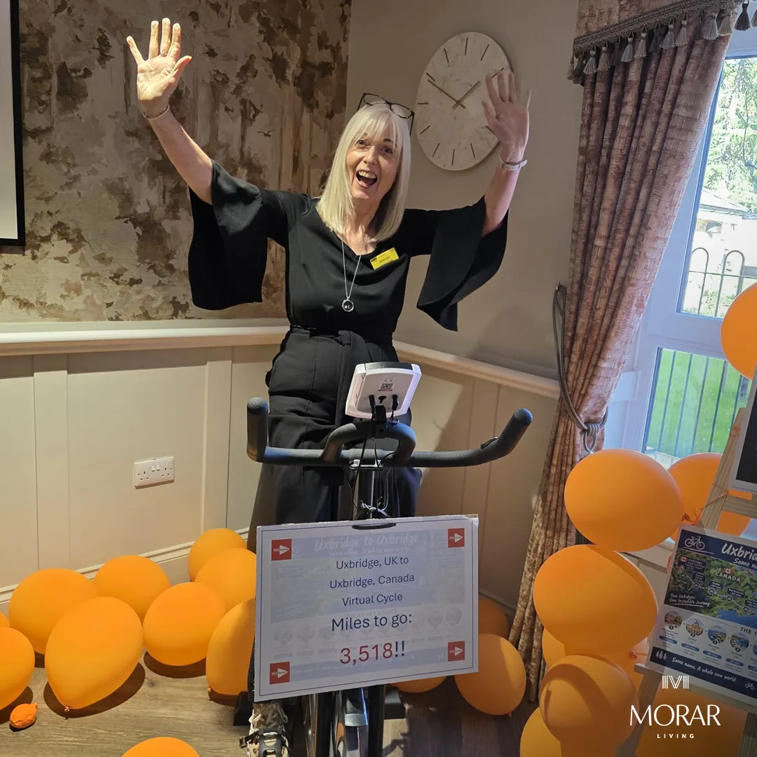A woman with both arms raised beside the exercise bike, surrounded by orange balloons, with a sign reading Miles to go: 3,518