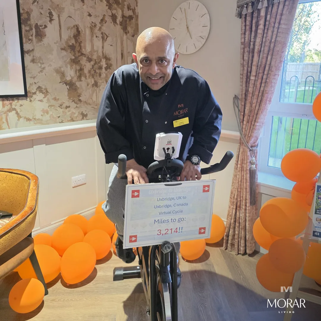 A man in a Morar chef's jacket riding the exercise bike, with a sign reading Miles to go: 3,214