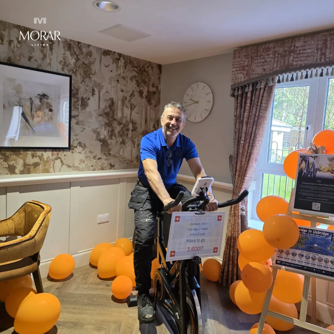 A smiling man in a blue polo shirt on the exercise bike in a lounge filled with orange balloons, with the route poster on an easel by the window