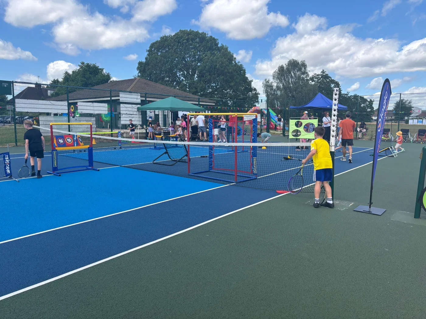 Children holding tennis rackets beside mini tennis nets on a blue court, with gazebos and families in the background