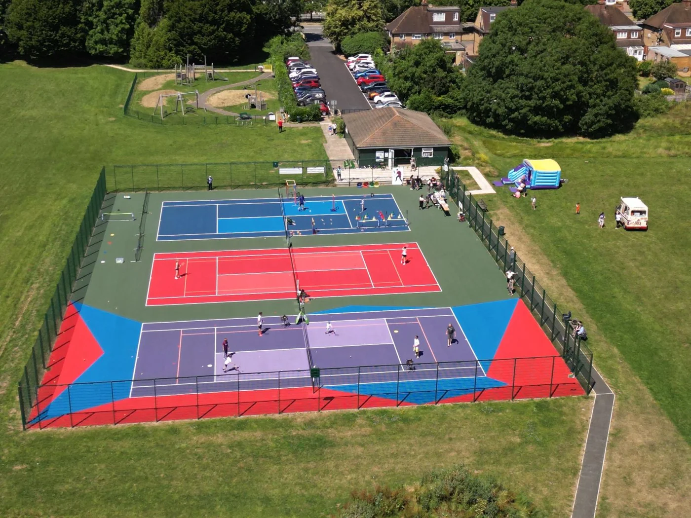 Aerial view of three outdoor tennis courts in use, with a pavilion, car park, playground and bouncy castle beside them