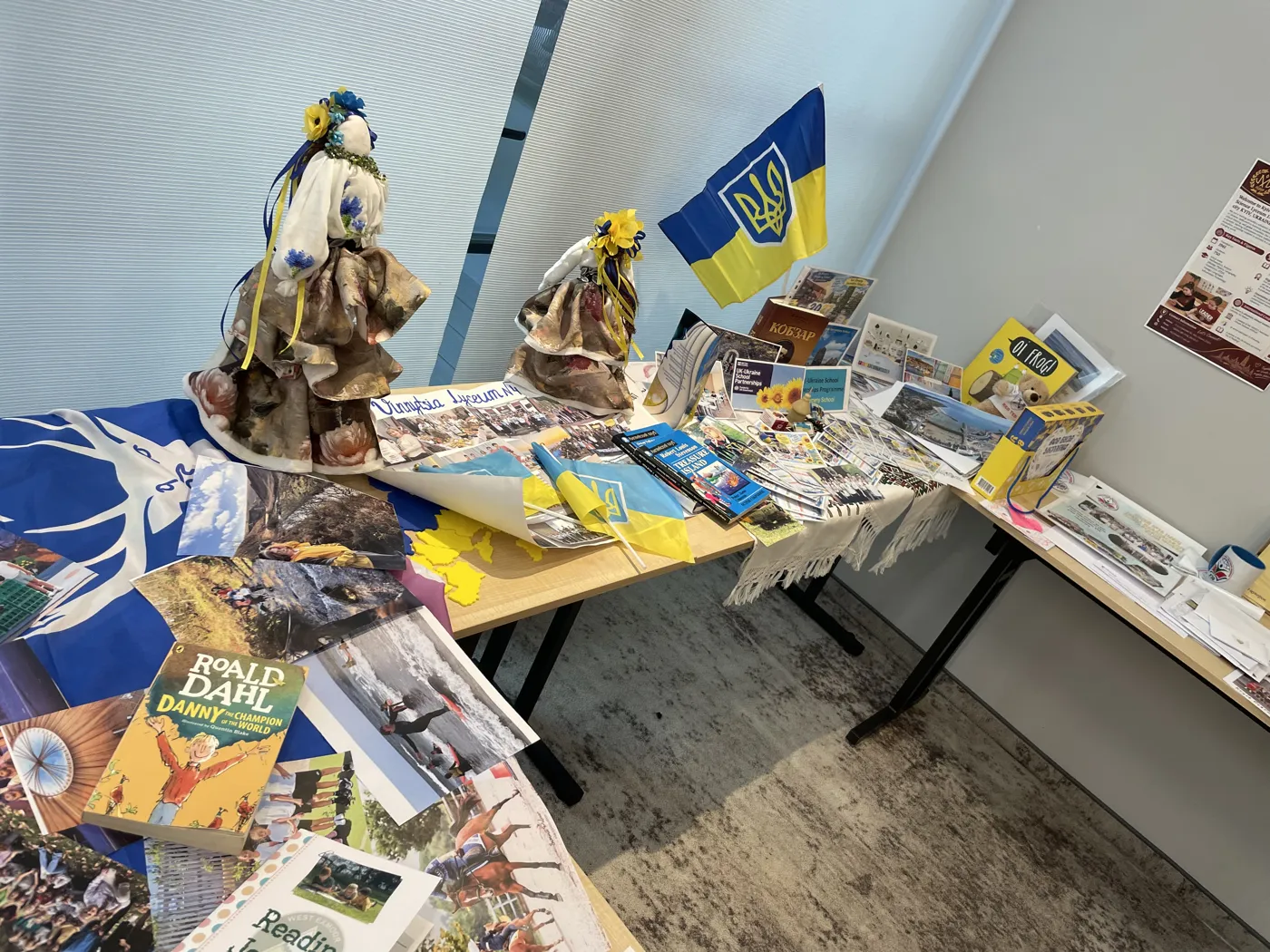 A long display table of books and crafts beneath a wall of pinned photographs, with a Union flag hanging from the table