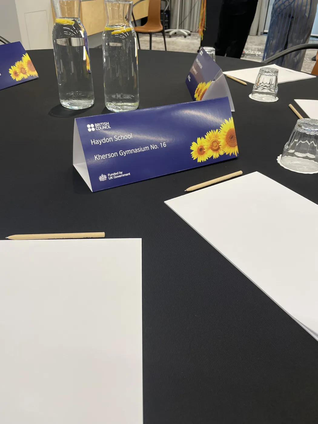 A British Council table card reading Haydon School and Kherson Gymnasium No. 16, decorated with sunflowers, on a conference table with water carafes and paper