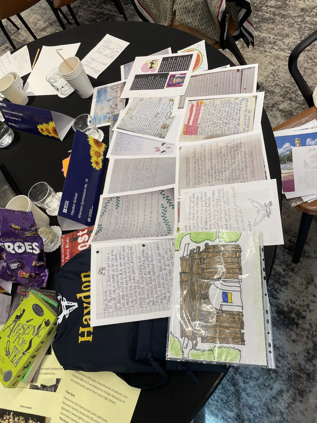 Handwritten letters and a child's drawing of a wooden house with a Ukrainian flag laid out on a conference table beside a navy Haydon School bag