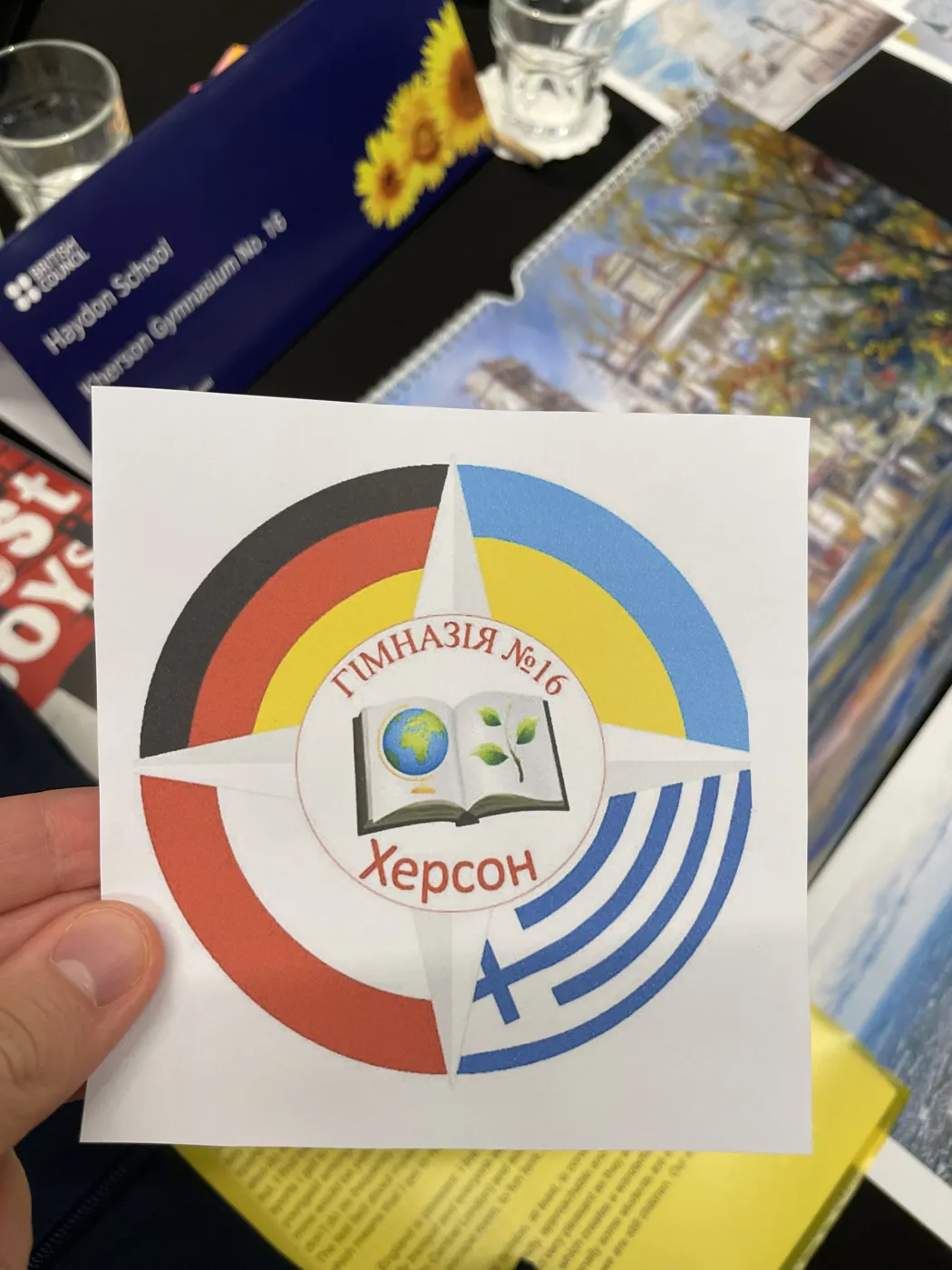 A hand holding a round sticker printed with the badge of Gymnasium No. 16 in Kherson, in Ukrainian, ringed with the colours of several national flags