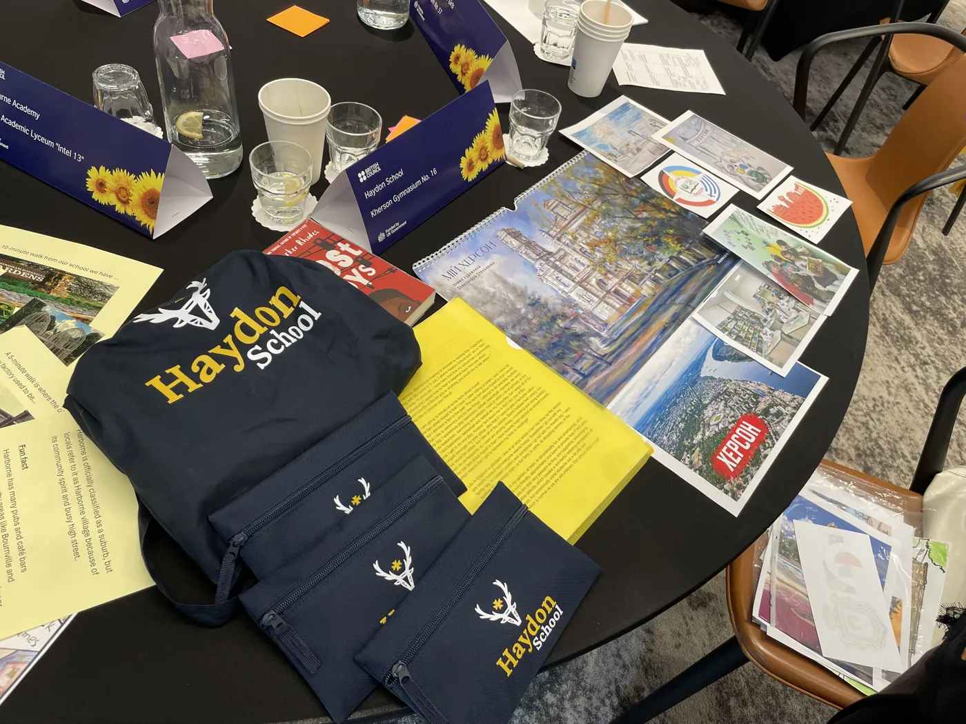 Navy Haydon School bags, printed postcards of Kherson and colourful papers spread across a round table