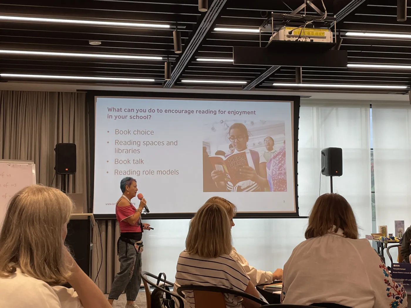 A speaker presenting a slide headed What can you do to encourage reading for enjoyment in your school, listing book choice, reading spaces and libraries, book talk and reading role models