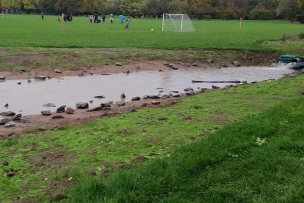 A shallow flood water storage basin holding water along the edge of the playing field at Bessingby Park, Ruislip, with goalposts behind